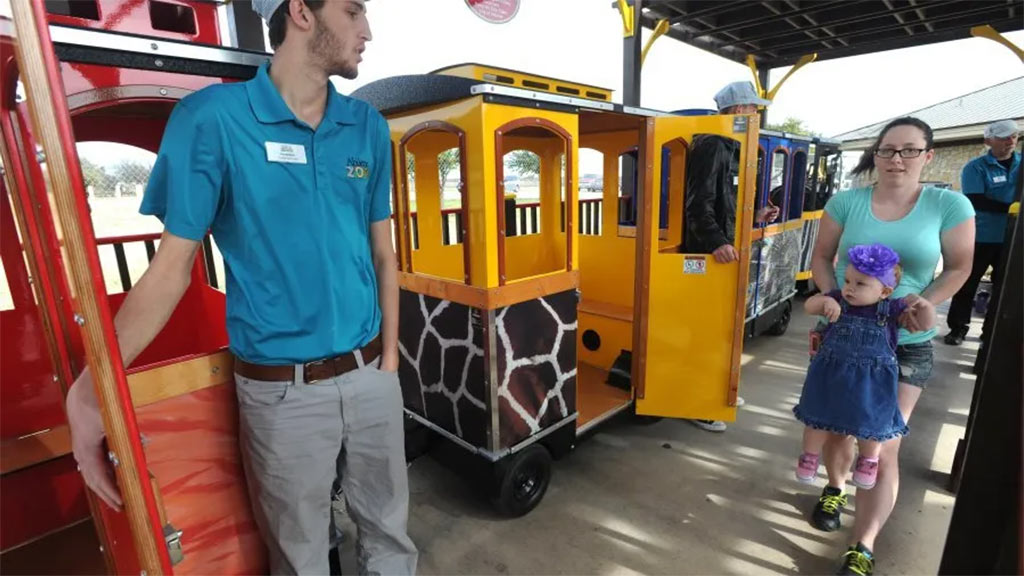 Zoo train driving past animal exhibits with children and parents on board in a busy zoo environment